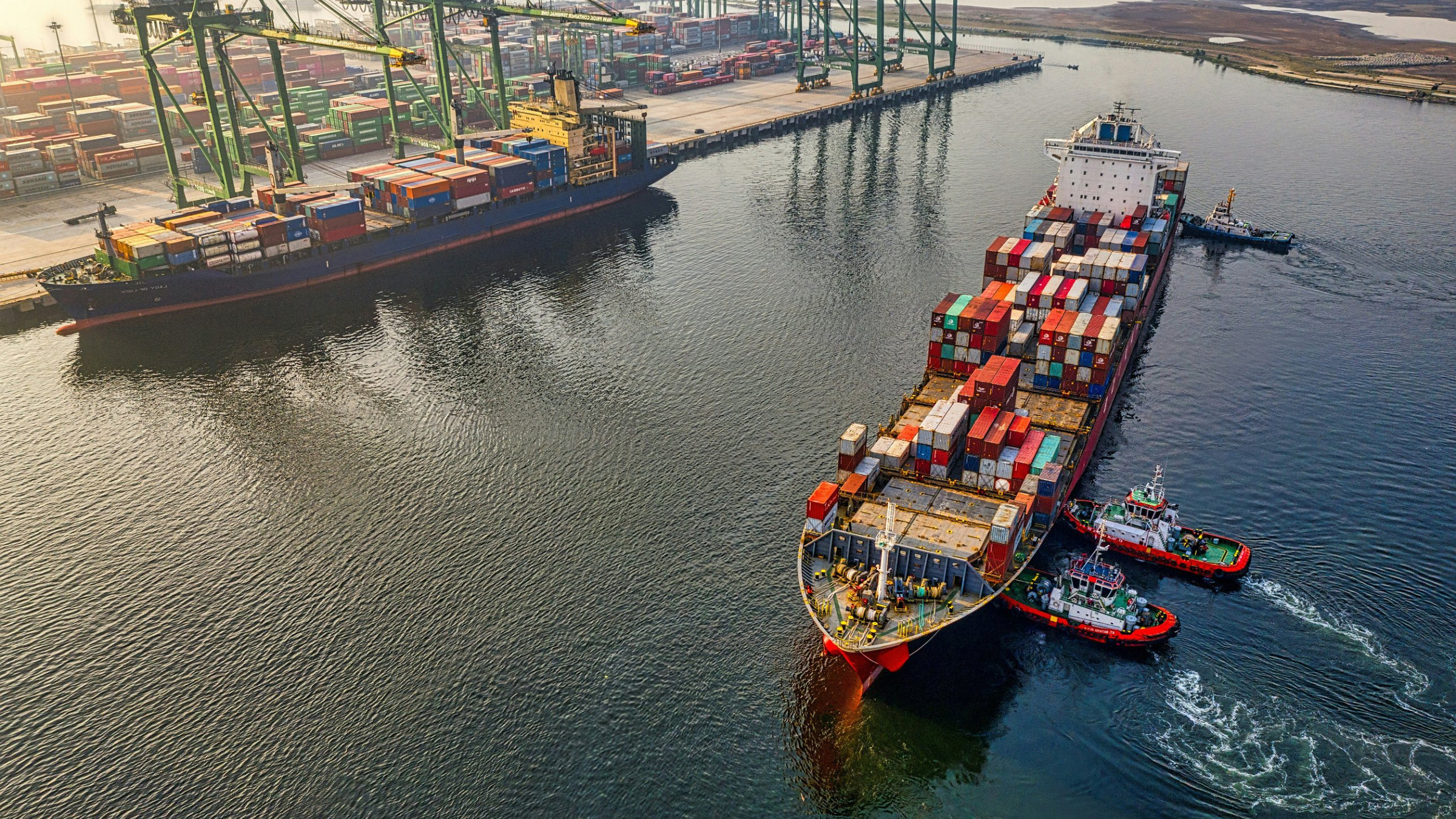 Aerial view of a container ship guided by tugboats beside a busy container terminal